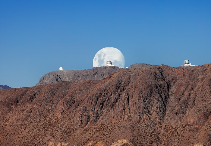 In a fantastically planned shot of the Moon, this image of Gemini South was timed to capture an almost perfectly full Moon framing the telescope. The other two telescopes caught between the red rocks of Cerro Pachón and the blue skies are the SOAR Telescope on the left and the almost finished Vera C. Rubin Observatory on the right. If you inspect the photograph closely, the red cranes being used to construct Rubin Observatory can just be spotted against the white dome. When complete, the telescope will be used to conduct an unprecedented, decade-long survey of the sky at optical wavelengths called the Legacy Survey of Space and Time (LSST). Gemini South is the southern member of the pair of telescopes that make up the international Gemini Observatory, a Program of NSF NOIRLab.