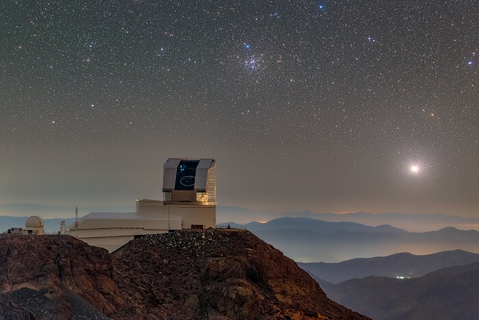 NSF–DOE Vera C. Rubin Observatory is seen with its dome open during First Look observation activities in April 2025. Messier 41, the Little Beehive Cluster, can be seen over the telescope in this telelens photo.